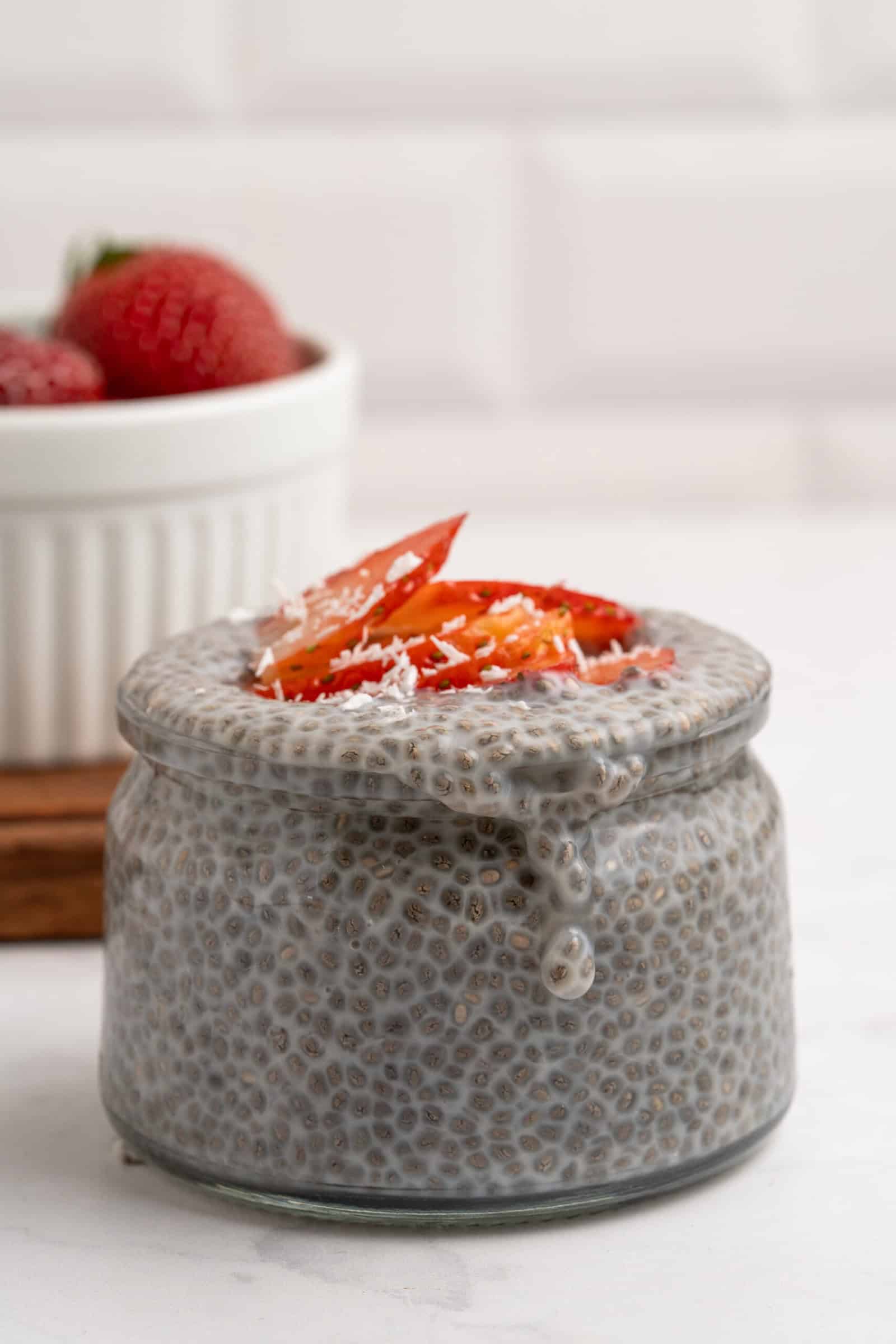 A glass jar filled with chia pudding is topped with sliced strawberries and shredded coconut. In the background, a white bowl holds whole strawberries. The scene is set on a white surface with a white tiled wall.