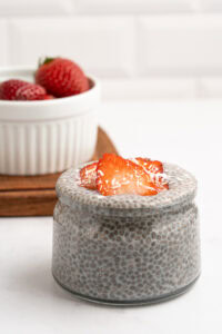 A small glass jar filled with chia pudding, topped with sliced strawberries and shredded coconut. In the background, a white bowl holds more strawberries on a wooden tray.