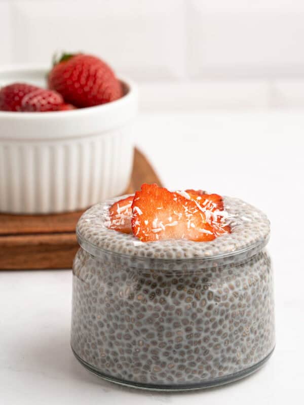 A small glass jar filled with chia pudding, topped with sliced strawberries and shredded coconut. In the background, a white bowl holds more strawberries on a wooden tray.