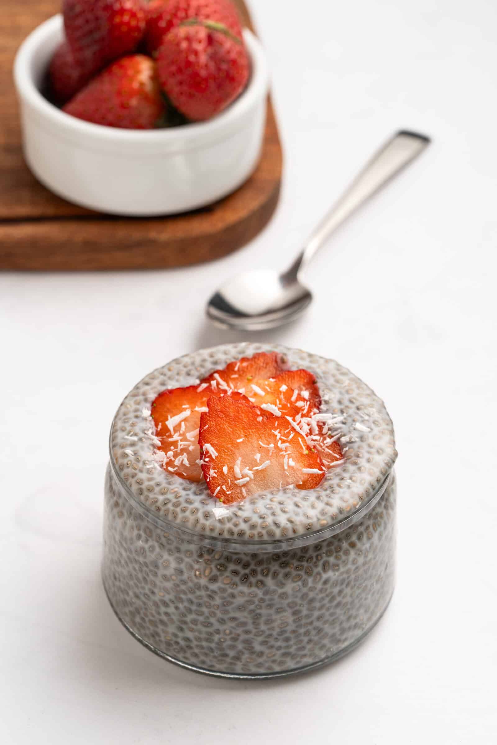 A glass jar of chia pudding topped with sliced strawberries and shredded coconut, with a spoon beside it and a bowl of whole strawberries in the background.