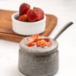 A glass jar filled with chia pudding, topped with sliced strawberries and shredded coconut, sits on a white surface. A spoon is inside the jar, and a small bowl of whole strawberries is in the background on a wooden board.