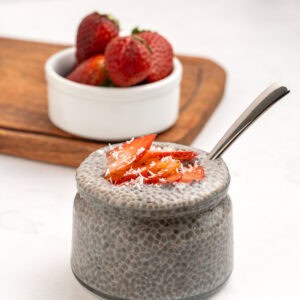 A glass jar filled with chia pudding, topped with sliced strawberries and shredded coconut, sits on a white surface. A spoon is inside the jar, and a small bowl of whole strawberries is in the background on a wooden board.