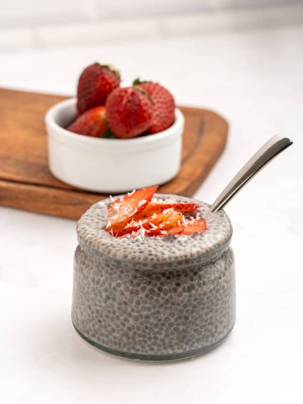 A glass jar filled with chia pudding, topped with sliced strawberries and shredded coconut, sits on a white surface. A spoon is inside the jar, and a small bowl of whole strawberries is in the background on a wooden board.