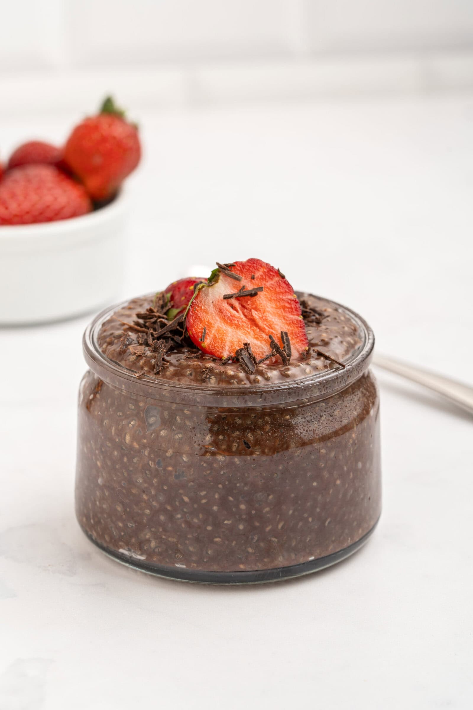 A small glass jar filled with chocolate chia pudding, topped with sliced strawberries and chocolate shavings. In the background, a white bowl of whole strawberries sits on a white surface.