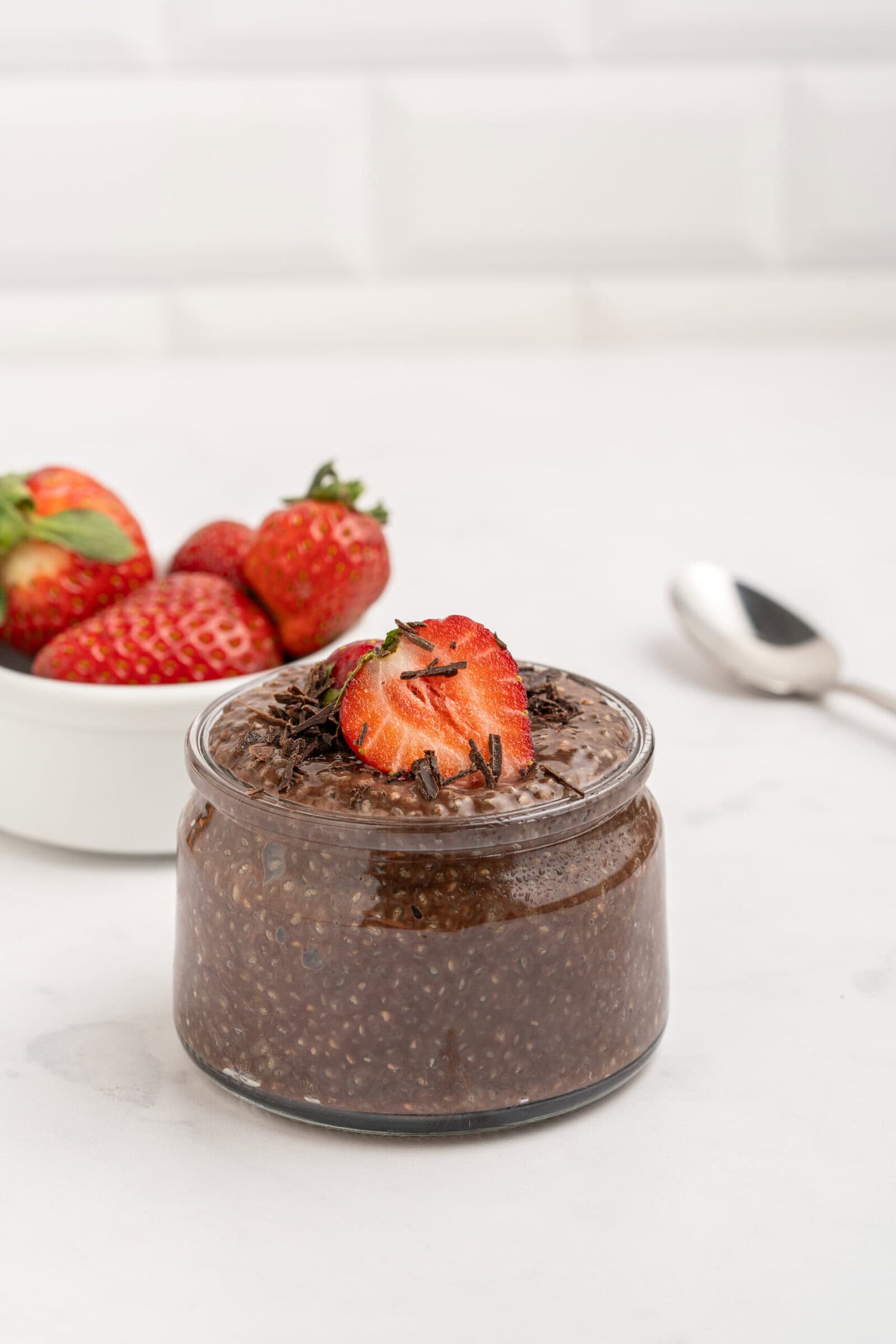 A glass jar of chocolate chia pudding topped with sliced strawberries and chocolate shavings sits on a white surface. In the background, there is a bowl of fresh strawberries and a spoon.