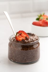 A glass jar filled with chocolate chia pudding, topped with a sliced strawberry and chocolate shavings, sits on a white surface. A spoon is in the jar, and a small bowl of strawberries is in the background.
