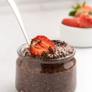 A glass jar filled with chocolate chia pudding, topped with a sliced strawberry and chocolate shavings, sits on a white surface. A spoon is in the jar, and a small bowl of strawberries is in the background.
