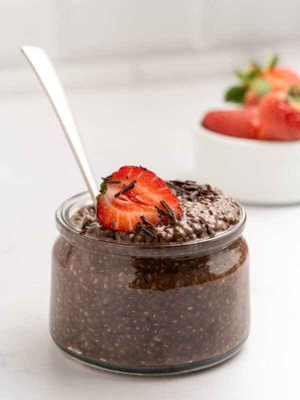 A glass jar filled with chocolate chia pudding, topped with a sliced strawberry and chocolate shavings, sits on a white surface. A spoon is in the jar, and a small bowl of strawberries is in the background.