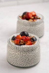 Two glass jars filled with chia pudding, topped with granola and fresh fruit pieces, including strawberries, grapes, and peach, placed on a white surface with a light background.