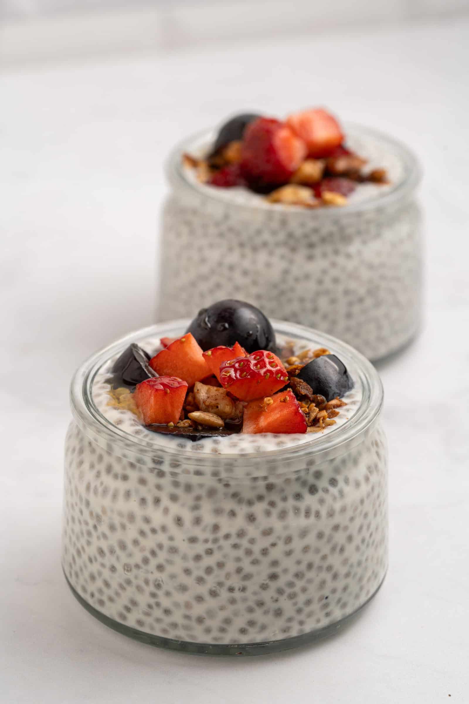Two glass jars filled with chia pudding, topped with granola and fresh fruit pieces, including strawberries, grapes, and peach, placed on a white surface with a light background.
