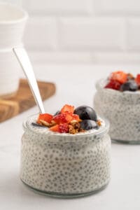 A glass jar filled with chia seed pudding, topped with granola, sliced strawberries, and blueberries. A spoon is placed in the jar, and another similar jar is visible in the background.