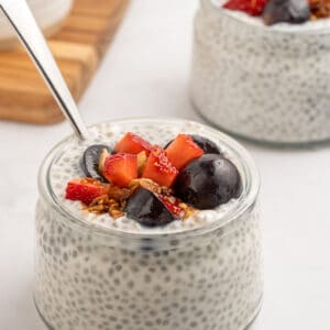 A small glass jar filled with chia pudding, topped with sliced strawberries, dark cherries, granola, and a spoon. Another similar jar is in the background on a white surface.