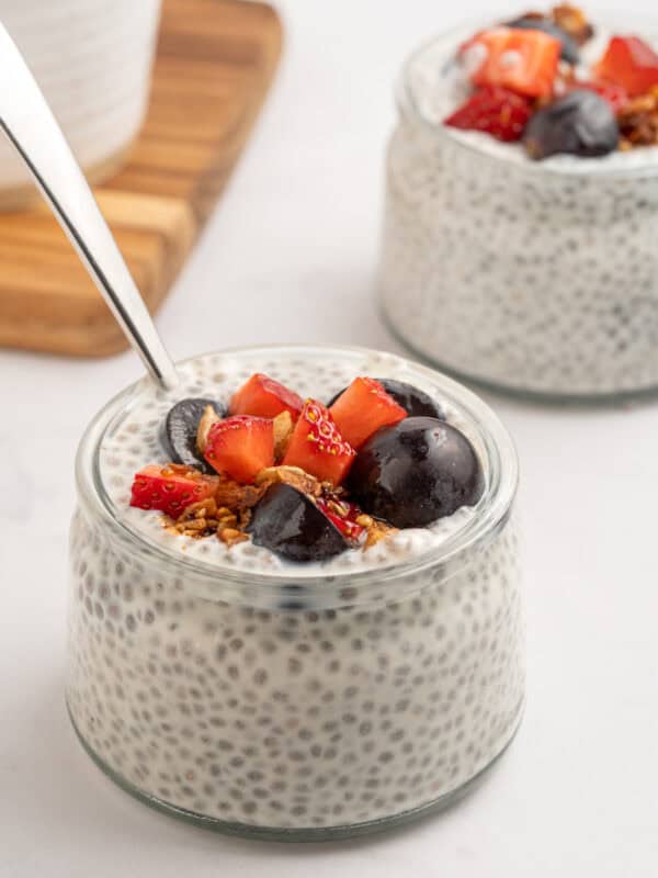 A small glass jar filled with chia pudding, topped with sliced strawberries, dark cherries, granola, and a spoon. Another similar jar is in the background on a white surface.