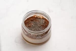A glass jar filled with layers of milk, cocoa powder, chia seeds, and nut butter sits on a white marble surface.