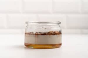 A glass jar on a white surface contains separated layers of honey, milk, cinnamon, and vanilla, against a white tiled background.