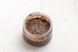 A small glass jar filled with a thick, dark brown, slightly textured mixture sits on a white, lightly marbled surface.