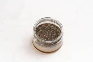 A small glass jar filled with chia seeds, partially submerged in a liquid, sits on a white surface.