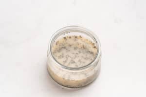 A small glass jar filled with a light-colored mixture containing chia seeds, sitting on a white, lightly textured surface.