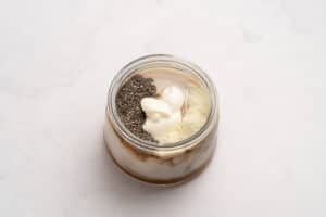 A glass jar filled with layers of yogurt, chia seeds, and protein powder, placed on a light marble surface.