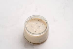 A small glass jar filled with creamy white sauce that has visible black pepper specks, placed on a light marble surface.