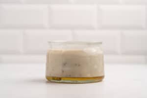 A glass jar filled with creamy beige sauce, possibly salad dressing, with visible herbs and a layer of oil at the bottom, set on a white surface with a white tiled background.