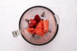 Top-down view of a blender containing ice cubes, frozen strawberries, and chunks of fresh watermelon on a light marble surface.
