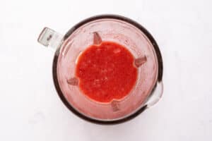 A top-down view of a blender containing a red, liquid mixture, likely a fruit smoothie or puree, on a white surface.