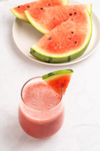 A glass of watermelon juice garnished with a small watermelon slice is on a white surface, with a plate of large watermelon wedges in the background.