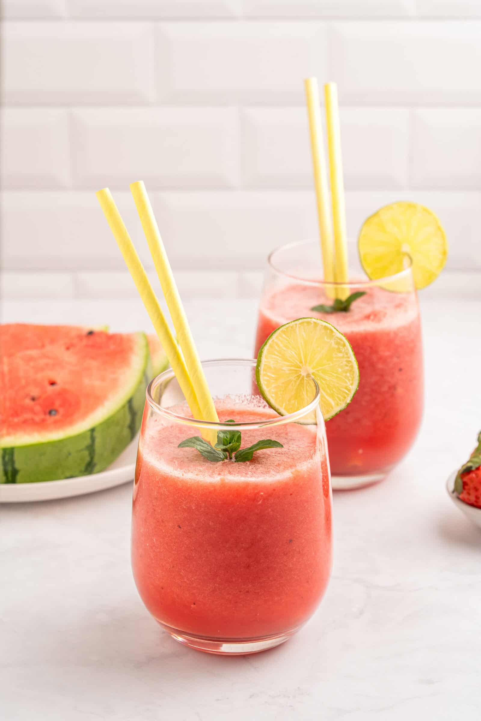 Two glasses of watermelon smoothie garnished with lime slices, mint leaves, and yellow straws. A slice of fresh watermelon is on a plate in the background, all set on a white surface with a tiled backdrop.
