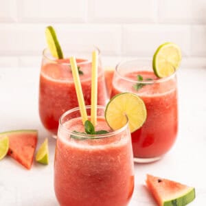 Three glasses of watermelon smoothie with lime slices and mint leaves as garnish, each with a yellow straw, surrounded by watermelon and lime wedges on a white surface.
