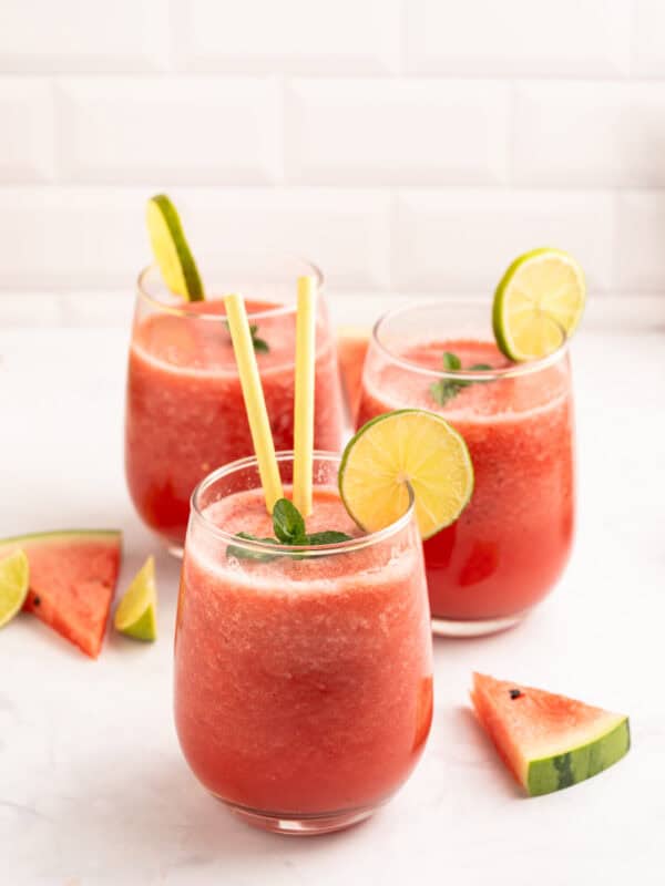 Three glasses of watermelon smoothie with lime slices and mint leaves as garnish, each with a yellow straw, surrounded by watermelon and lime wedges on a white surface.