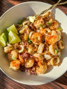 A white bowl filled with cooked shrimp and calamari, garnished with herbs and served with lime wedges, sits on a wooden surface in bright sunlight.