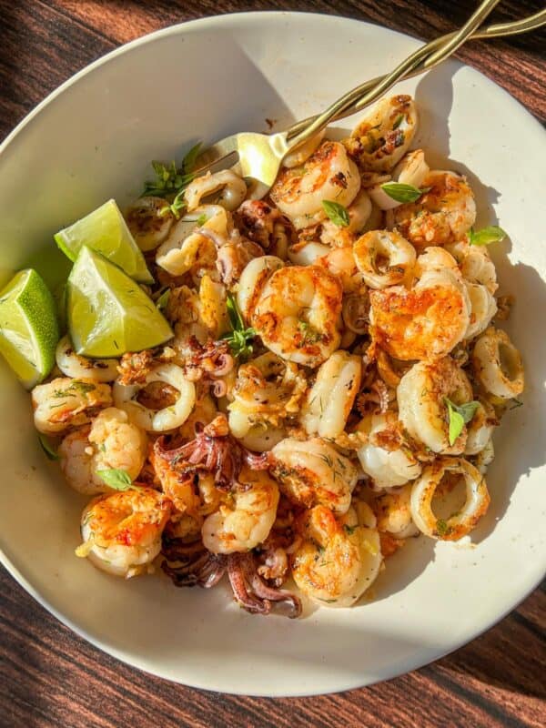 A white bowl filled with cooked shrimp and calamari, garnished with herbs and served with lime wedges, sits on a wooden surface in bright sunlight.