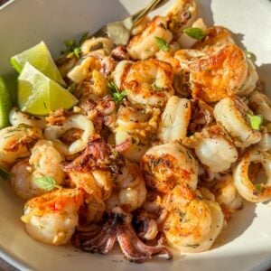 A white bowl filled with grilled shrimp and calamari, garnished with herbs and served with lime wedges, sits on a wooden surface in bright natural light.