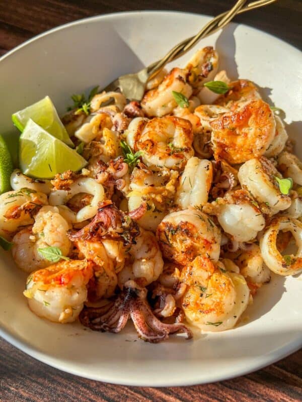 A white bowl filled with grilled shrimp and calamari, garnished with herbs and served with lime wedges, sits on a wooden surface in bright natural light.