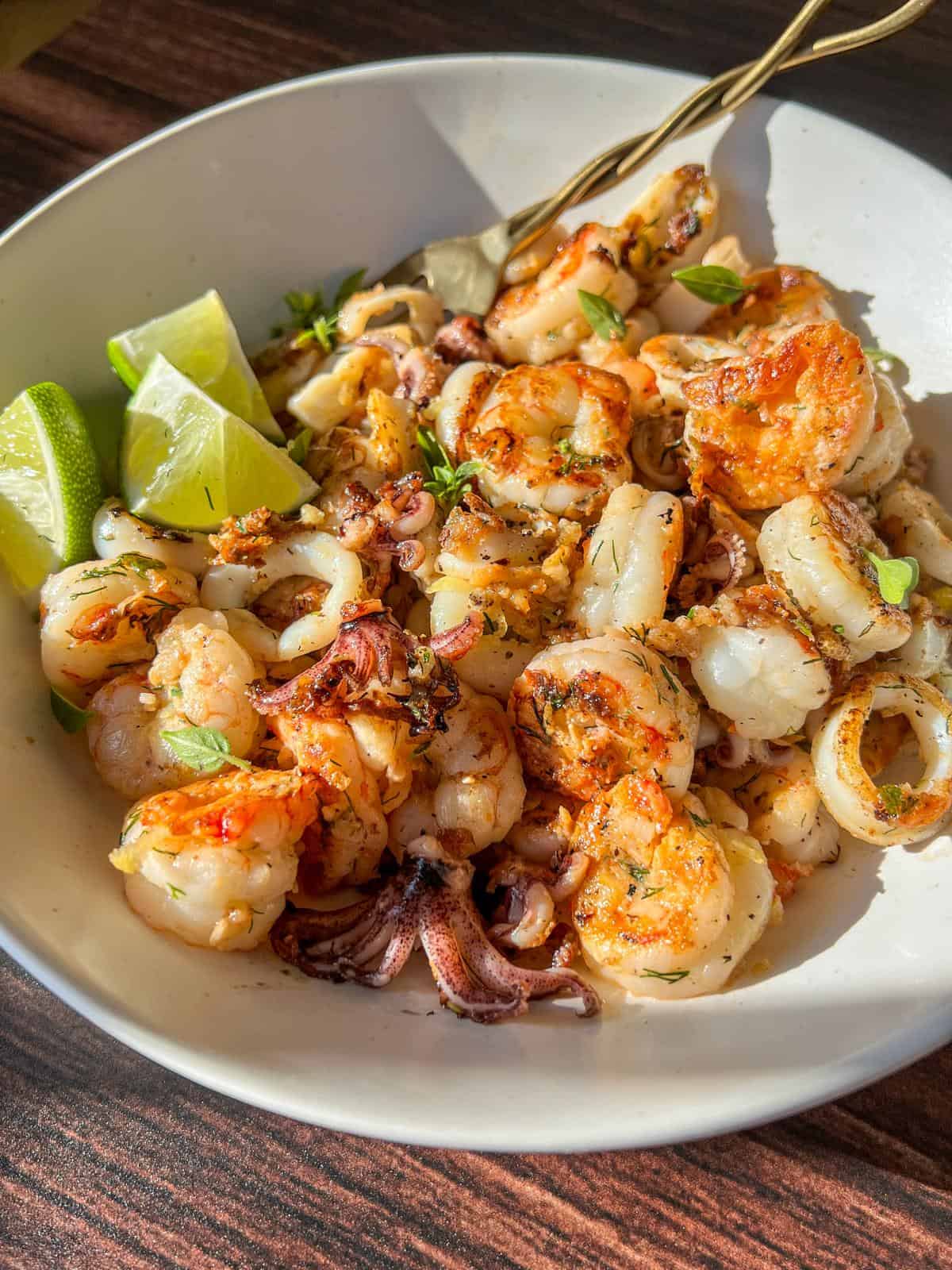 A bowl of grilled seafood, including shrimp and squid, garnished with fresh herbs and served with lime wedges, sits on a wooden table in bright sunlight.