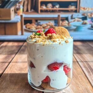 A glass filled with creamy dessert layered with raspberries, cookies, and crumbs, topped with raspberries and mint, sits on a wooden table with a bakery display blurred in the background.