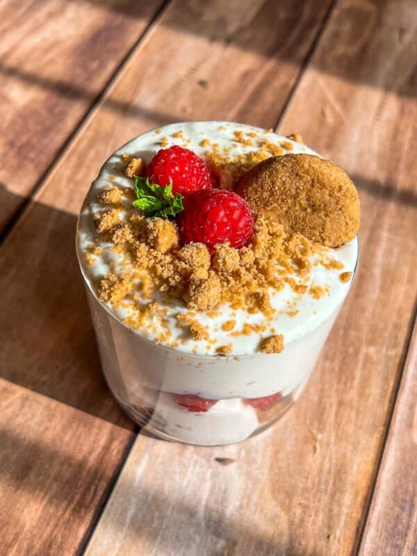 A dessert cup with creamy layers, topped with fresh raspberries, crumbled cookie, a whole cookie, and a small mint leaf, placed on a wooden surface with sunlight casting shadows.