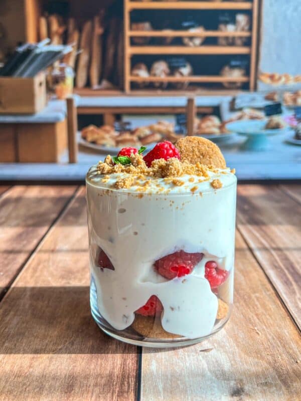 A glass filled with creamy dessert layered with raspberries, cookies, and crumbs, topped with raspberries and mint, sits on a wooden table with a bakery display blurred in the background.