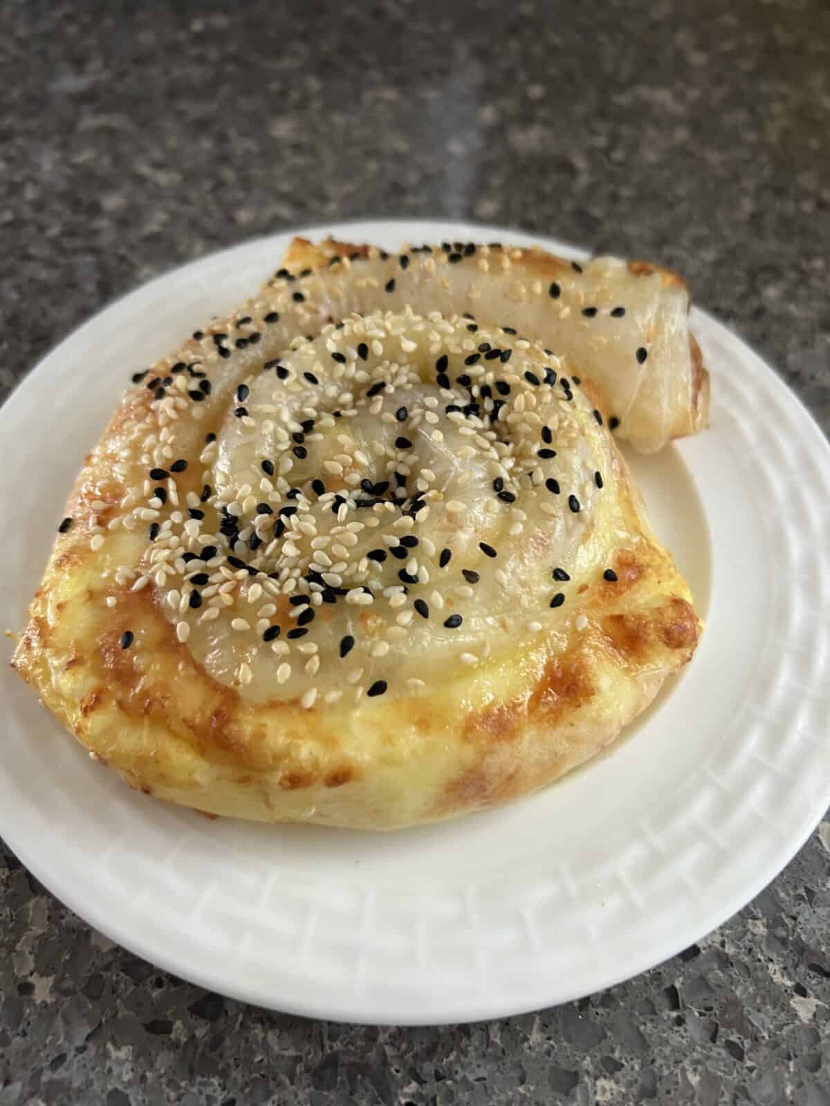 A savory pastry roll topped with melted cheese, black and white sesame seeds, served on a white plate over a granite countertop.