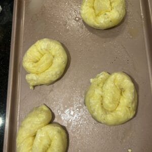 Four unbaked, twisted dough knots—reminiscent of Rice Paper Burek—are brushed with egg wash and rest on a metal baking sheet, ready to be baked.