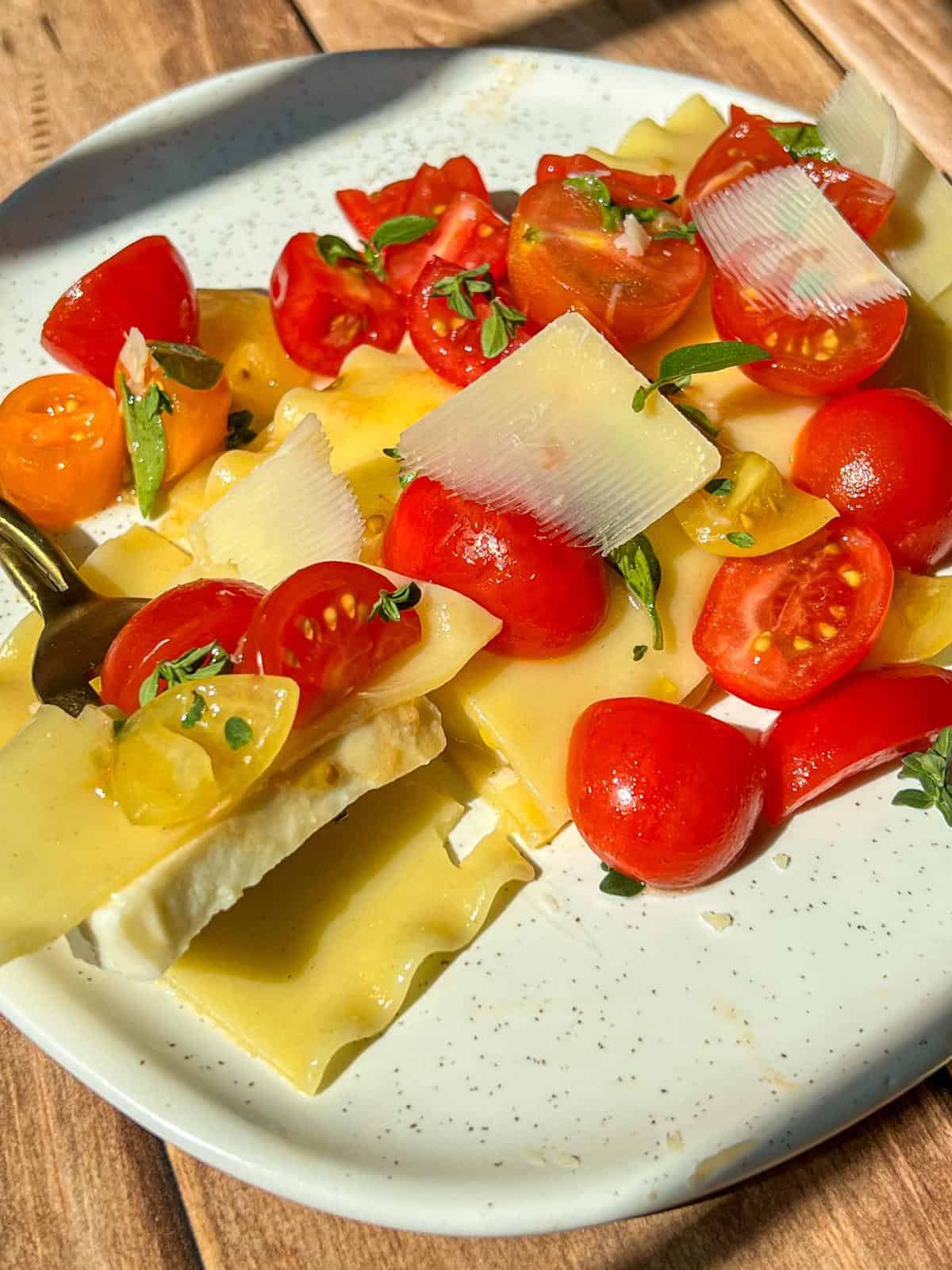A plate of lasagna sheets topped with halved cherry tomatoes, fresh herbs, and thin cheese slices, all drizzled with olive oil. A fork holds a bite of lasagna, tomatoes, and cheese. Sunlight brightens the dish.