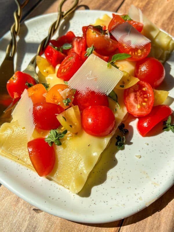 A close-up of lasagna noodles topped with cherry tomatoes, yellow tomatoes, fresh basil, and shaved cheese on a white plate, with two forks beside the food. Sunlight casts bright shadows on the wooden table.