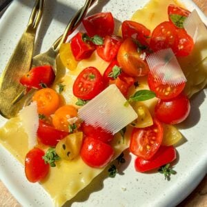 A plate with lasagna sheets topped with cherry tomato halves, fresh herbs, and thin slices of cheese. A gold fork and knife rest on the side, and sunlight casts shadows on the wooden table.