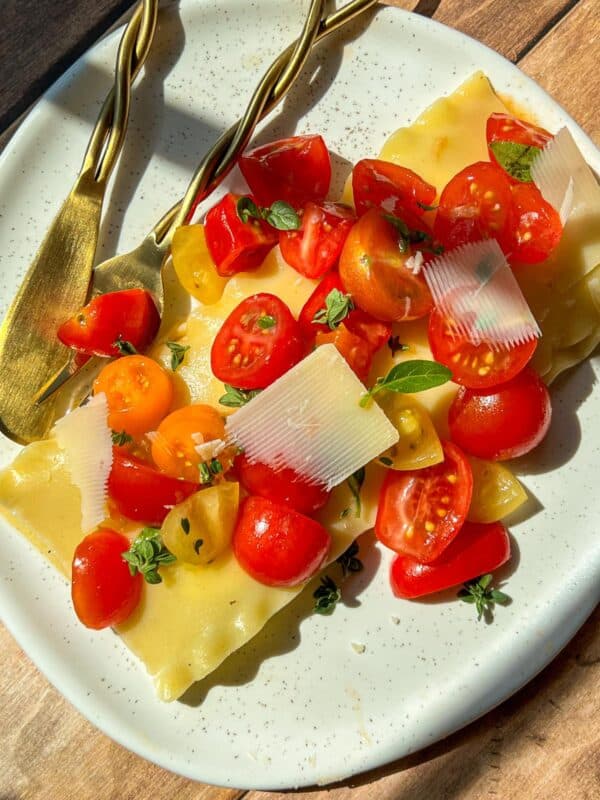 A plate with lasagna sheets topped with cherry tomato halves, fresh herbs, and thin slices of cheese. A gold fork and knife rest on the side, and sunlight casts shadows on the wooden table.