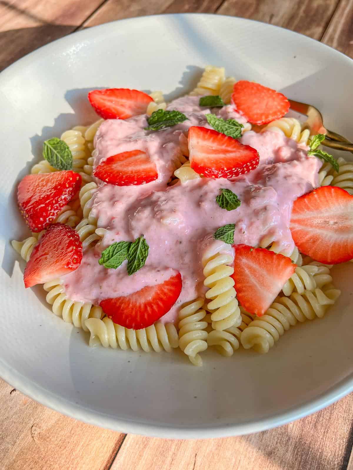 A white plate of fusilli pasta topped with pink strawberry sauce, fresh strawberry slices, and mint leaves, set on a wooden surface in natural light.