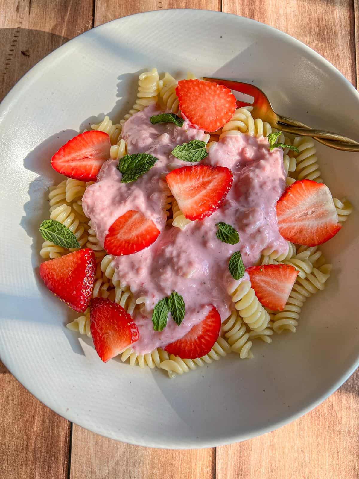 A white bowl with spiral pasta topped with pink strawberry cream sauce, fresh strawberry slices, and mint leaves, set on a wooden surface with a gold fork.