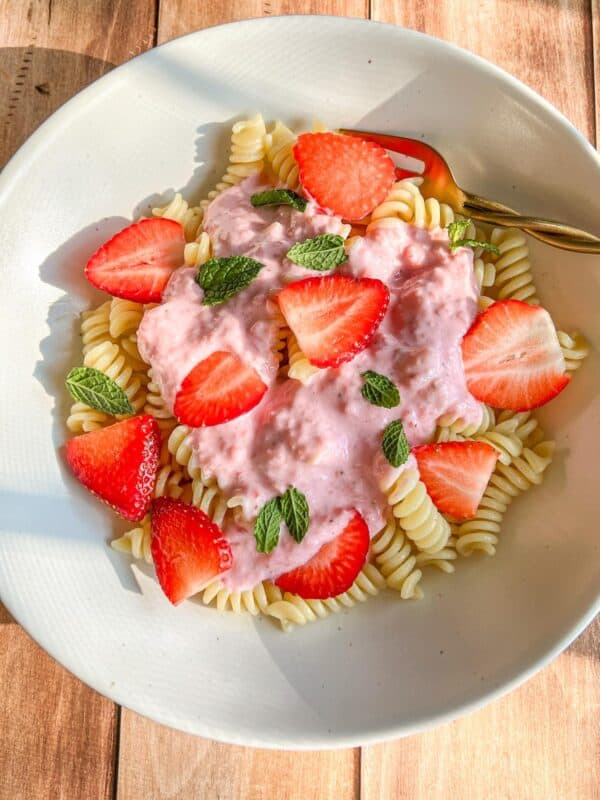 A white bowl of rotini pasta topped with a creamy pink strawberry sauce, fresh strawberry slices, and mint leaves, with a gold fork on the side, on a wooden surface.