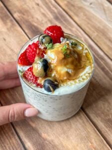 A hand holds a glass of chia pudding topped with strawberries, blueberries, gooseberries, a dollop of peanut butter, honey, and a sprig of fresh herbs, set on a wooden surface.
