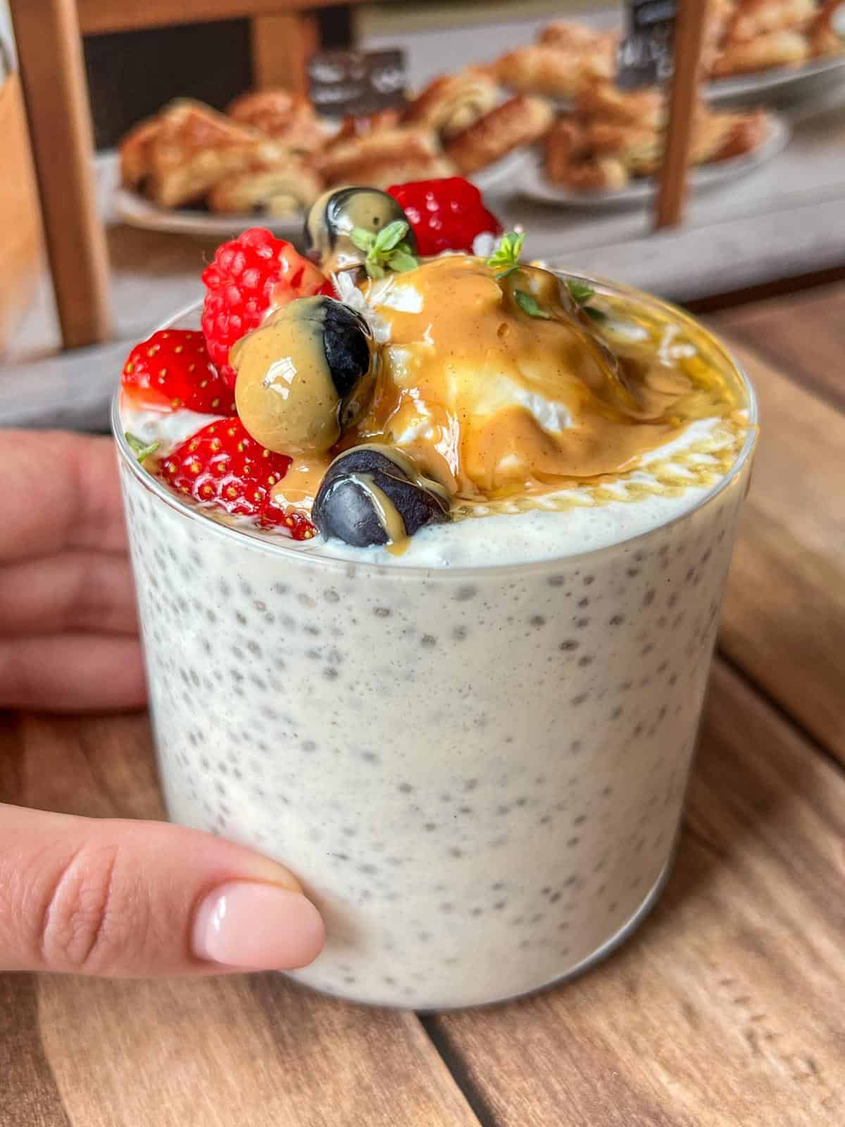 A glass of chia pudding topped with fresh strawberries, blueberries, a golden berry, and a generous dollop of peanut butter. A hand is holding the glass on a wooden table, with pastries blurred in the background.